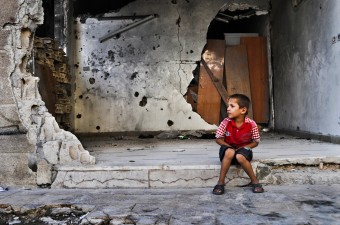 Criança na entrada de sua casa destruída em Holms. Foto: PMA / Abeer Etefa